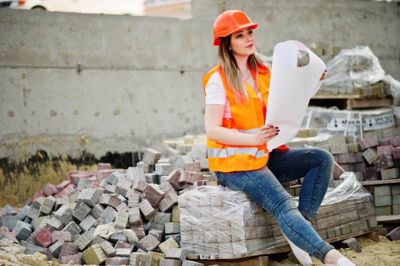 Engineer builder woman in uniform waistcoat and orange protective helmet hold business layout plan paper sitting on pavement.